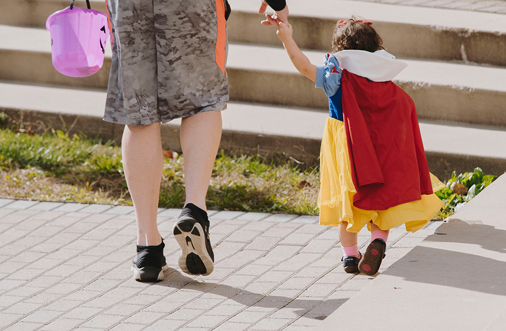 A guardian holding a child's hand, child is wearing costume and adult is holding a pumpkin-shaped candy bucket; walking